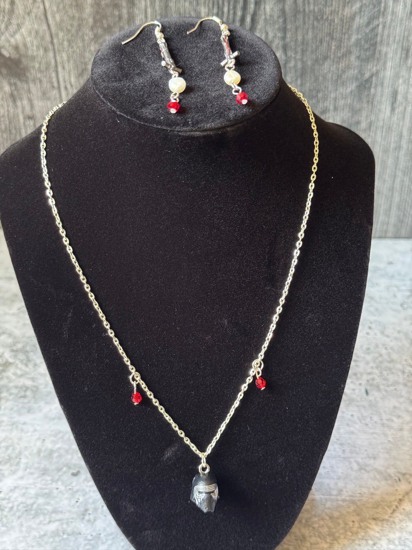 Shows a silver chain necklace with red bead charms and a black helmet from a toy and earrings made from toy lightsaber hilts with pearl and red bead charms on bottom. 