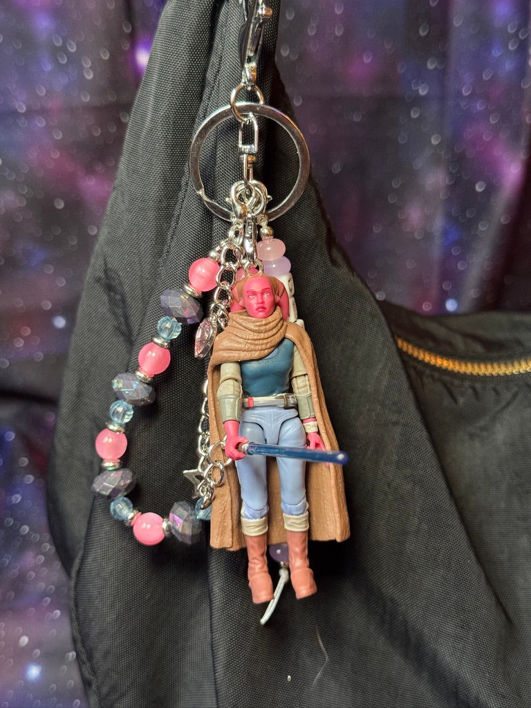 Devon Izara character from Star Wars action figure that has been turned into a bag charm/keychain. Attached to a silver keychain with coordinating beads and charms in a pink, blue and silver color scheme. Bag charm is attached to a black purse set against a purple background. 