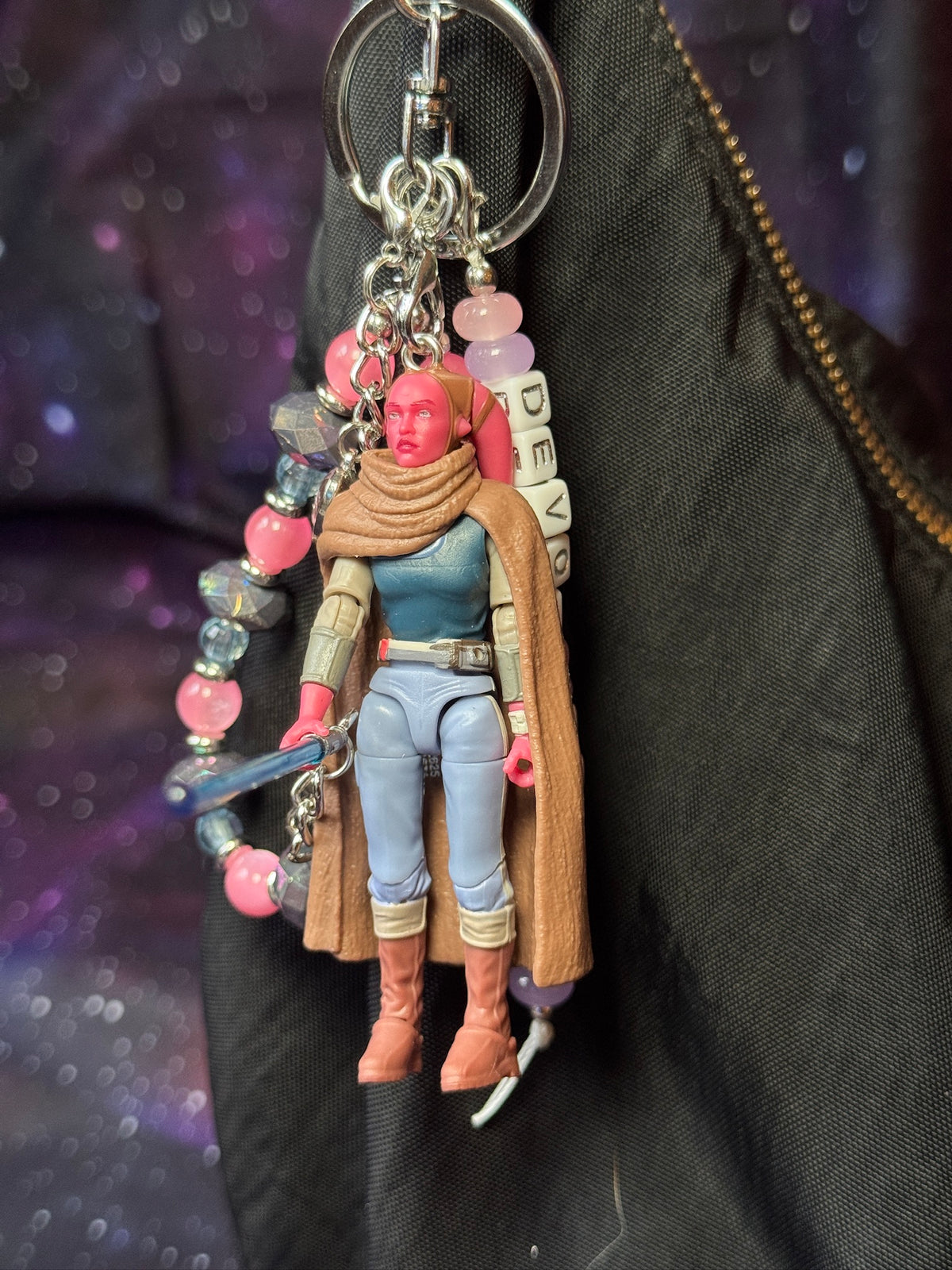 Devon Izara character from Star Wars action figure that has been turned into a bag charm/keychain. Attached to a silver keychain with coordinating beads and charms in a pink, blue and silver color scheme. Bag charm is attached to a black purse set against a purple background. 