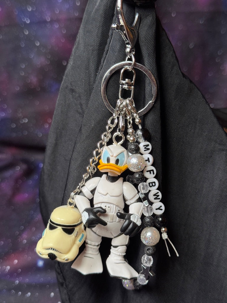 Donald Duck styled as a Stormtrooper from Star Wars figure. Attached to a silver keychain with coordinating beads in a black, silver and white color scheme. Figure is attached to a black purse set against a purple background. 