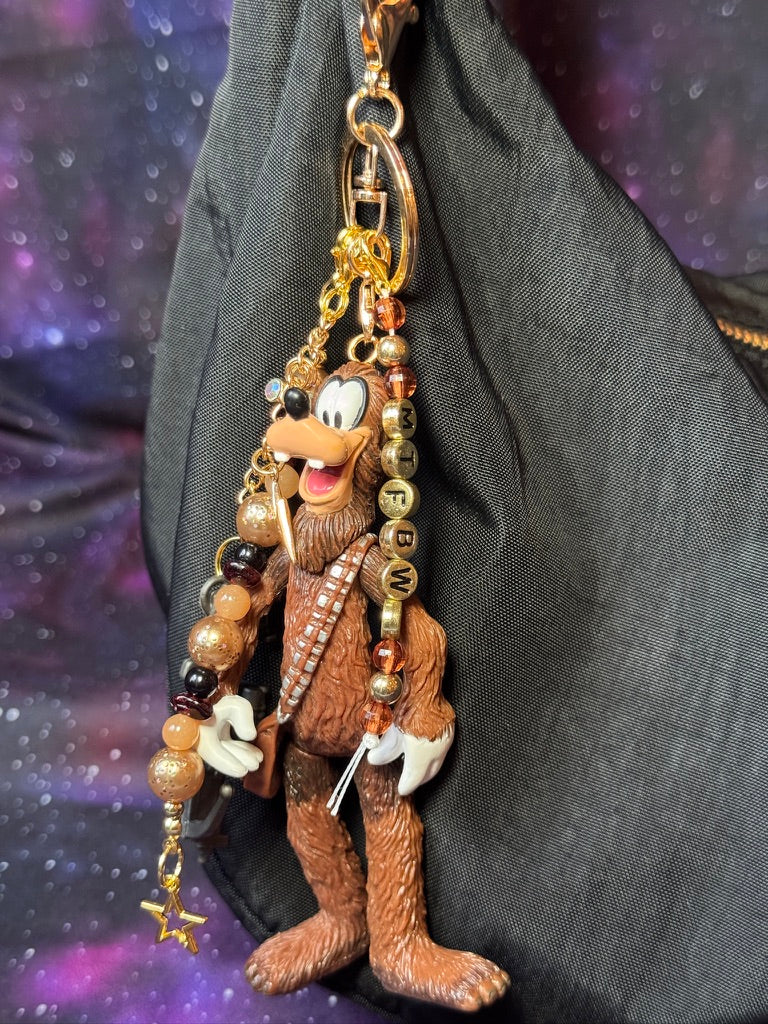 Goofy styled as Chewbacca from Star Wars figure. Attached to a gold keychain with coordinating beads in a brown gold and black color scheme. Figure is attached to a black purse set against a. purple background. 