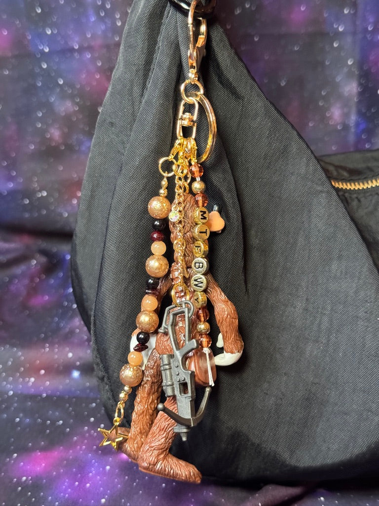 Goofy styled as Chewbacca from Star Wars figure. Attached to a gold keychain with coordinating beads in a brown gold and black color scheme. Figure is attached to a black purse set against a. purple background. 
