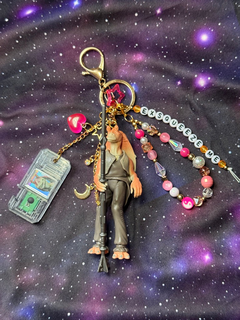 Jar Jar Binks character from Star Wars action figure that has been turned into a bag charm/keychain. Attached to a gold keychain with coordinating beads and charms in a pink, orange and gold color scheme. Figure is set against a purple background. Letter beads say "Exsqueeze Me."