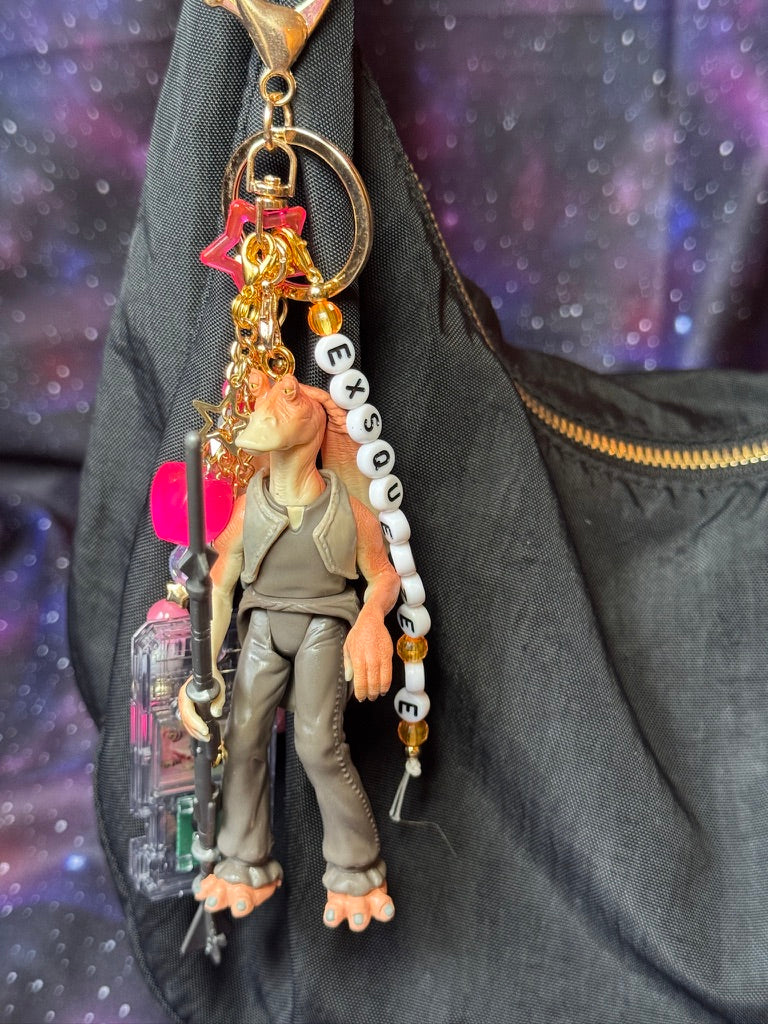Jar Jar Binks character from Star Wars action figure that has been turned into a bag charm/keychain. Attached to a gold keychain with coordinating beads and charms in a pink, orange and gold color scheme. Figure is attached to a black purse set against a purple background. 