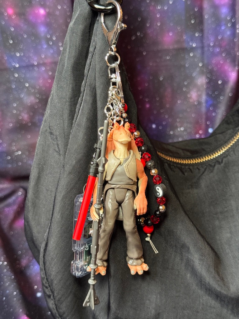 Jar Jar Binks character from Star Wars action figure that has been turned into a bag charm/keychain. Attached to a silver keychain with coordinating beads and charms in a black, silver and red color scheme. Figure is attached to a black purse set against a purple background. 