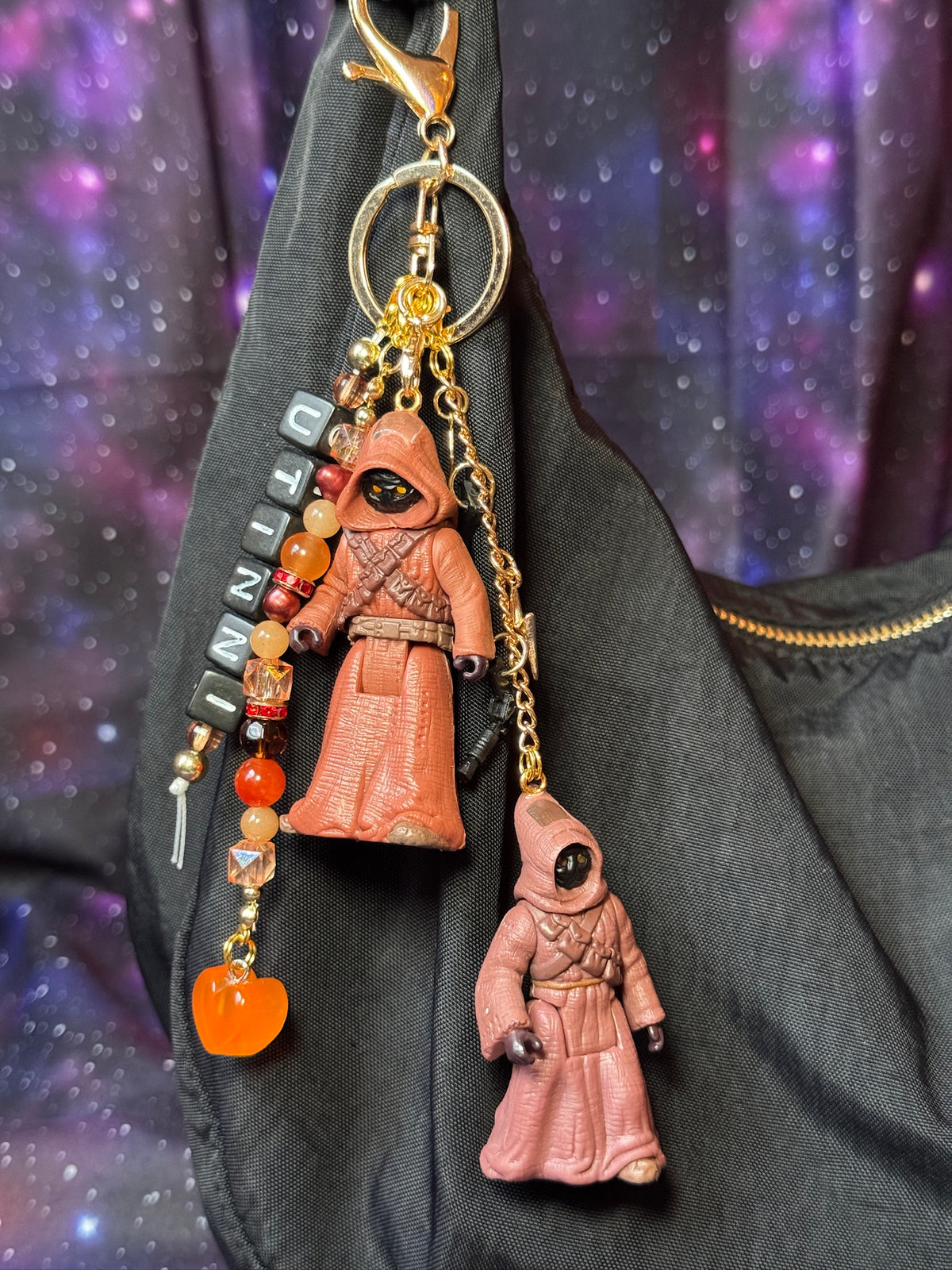 Jawas character from Star Wars action figures that have been turned into a bag charm/keychain. Attached to a gold keychain with coordinating beads and charms in a brown, orange, black and gold color scheme. Bag charm is attached to a black purse set against a purple background. 