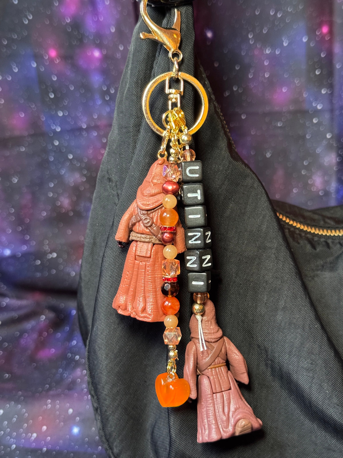 Jawas character from Star Wars action figures that have been turned into a bag charm/keychain. Attached to a gold keychain with coordinating beads and charms in a brown, orange, black and gold color scheme. Bag charm is attached to a black purse set against a purple background. 