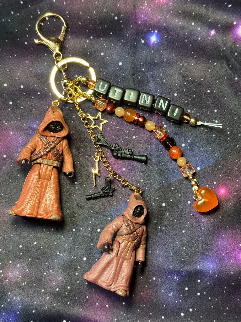 Jawas character from Star Wars action figures that have been turned into a bag charm/keychain. Attached to a gold keychain with coordinating beads and charms in a brown, orange, black and gold color scheme. Bag charm is set against a purple background. Message charm says "Utinni."